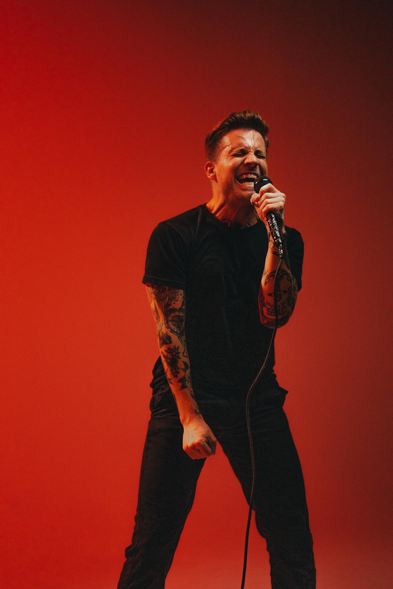Tattooed male performer sings energetically into a microphone against a bold red background.
